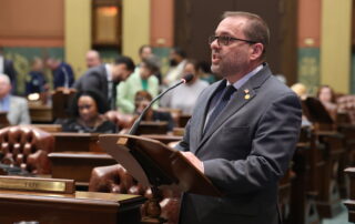 State Rep. Mike McFall speaks into a microphone at a podium on the Michigan House floor.