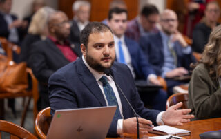 Rep. Farhat testifies at a table in front of a microphone in committee. Laptop open at left.