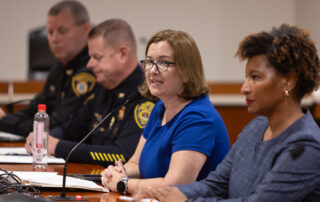 State Rep. Natalie Price testifies on her House Bill 5696 into a microphone at a table while surrounded by local officials.