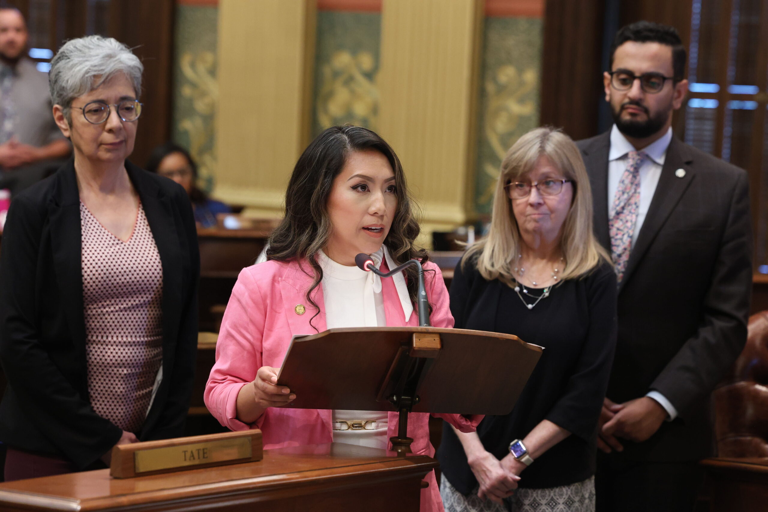 Michigan state Representative Mai Xiong stands at a podium between her colleagues, speaking on the House Floor in the Michigan Capitol Building.
