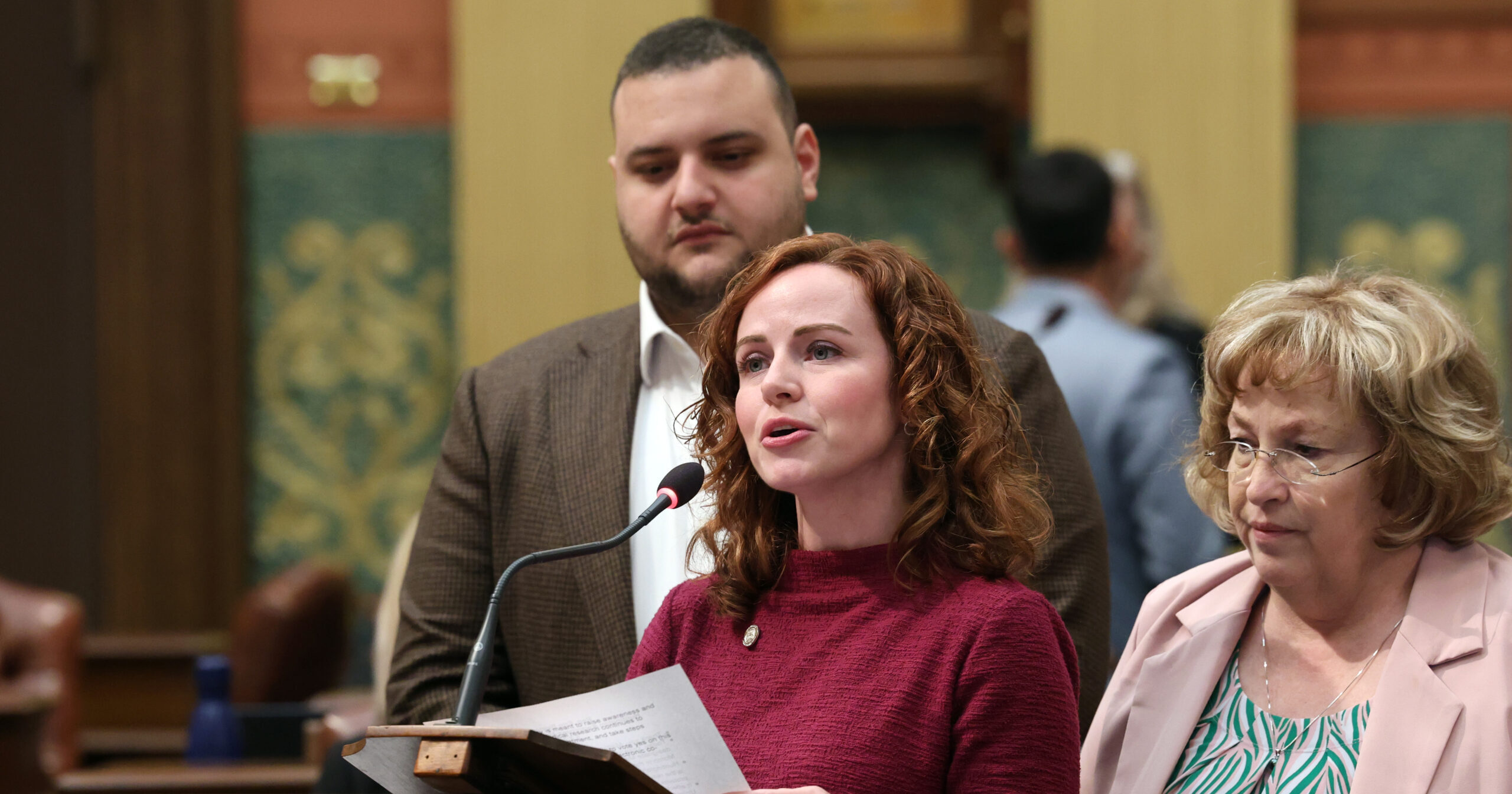 Michigan state Representative Erin Byrnes giving a speech on the House Floor supported by state Reps. Reggie Miller and Alabas Farhat.