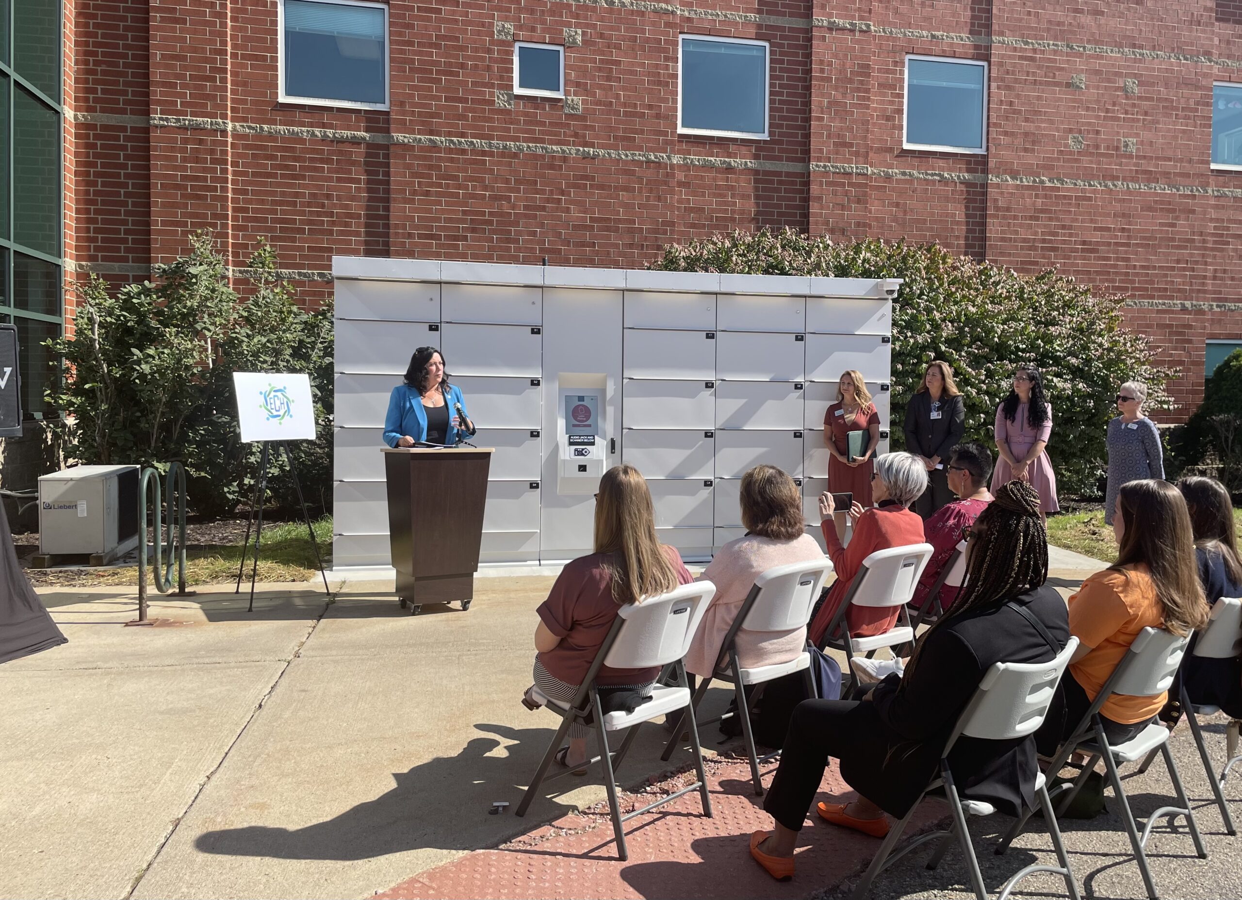 Representative Angela Witwer speaks at a podium in front of Michigan's first smart food lockers to a crowd of attendees at the ribbon-cutting ceremony in Eaton County.