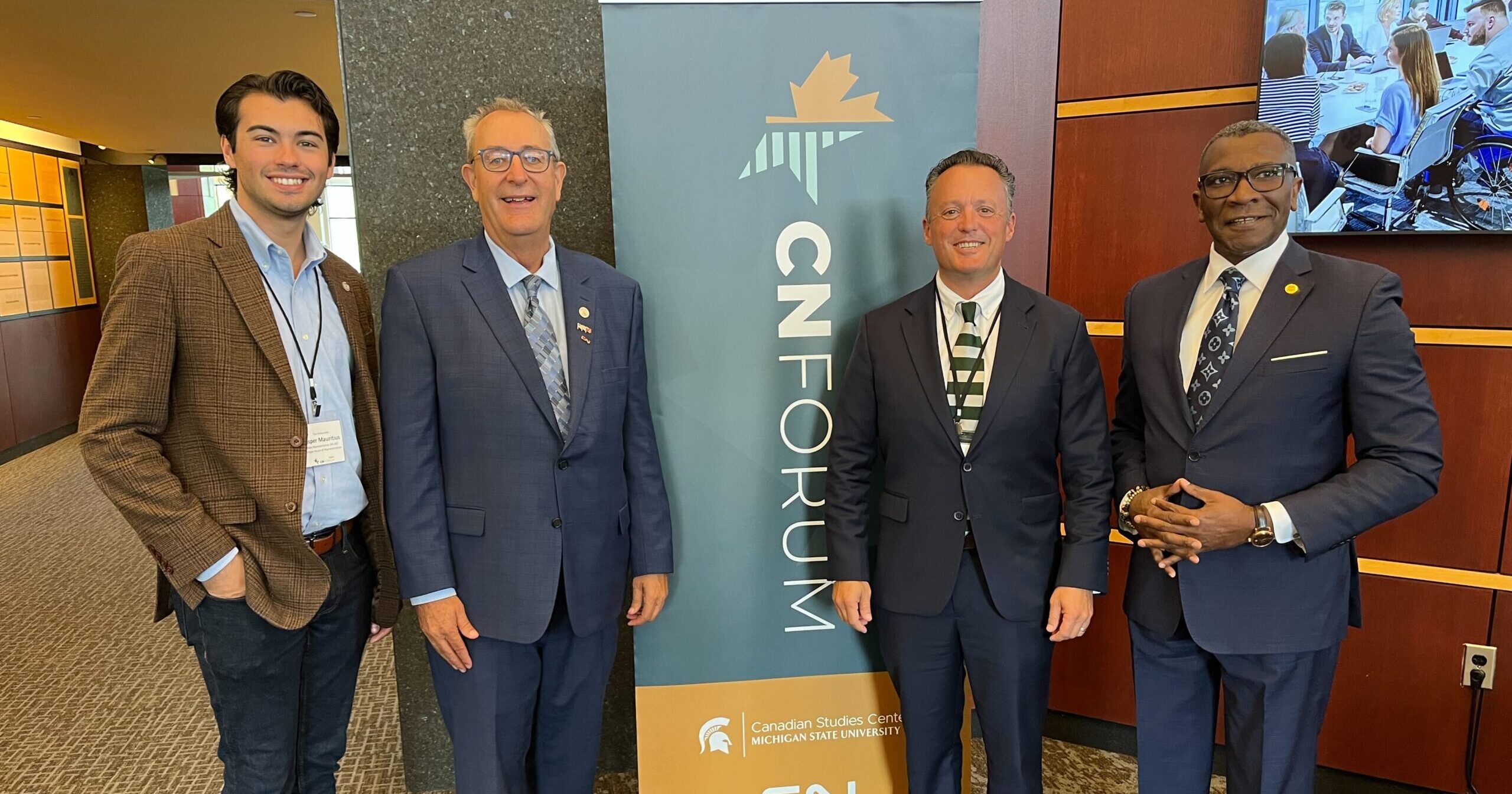 From left to right, Michigan state Representive Jasper R. Martus, state Sen. Roger Victory, President of the Michigan Railroad Association, Jon Cool and state Rep. Amos O’Neal stand smiling at the conference in front of a green and yellow poster.