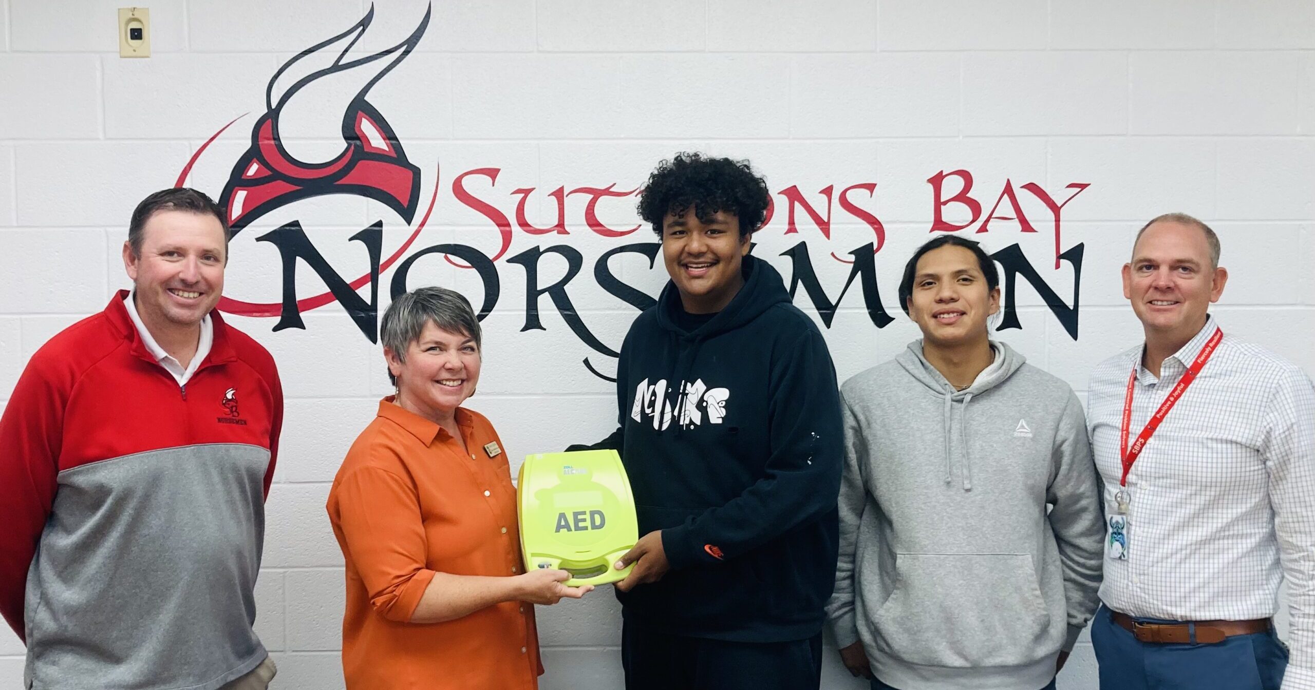 Michigan State Representative Betsy Coffia passing an automated external defibrillator to students Zachariah Raphael, Damien Chippewa, Superintendent Casey Petz and Chief Operations Officer Tim Smith in front of a Suttons Bay Norsemen mural at Suttons Bay High School.