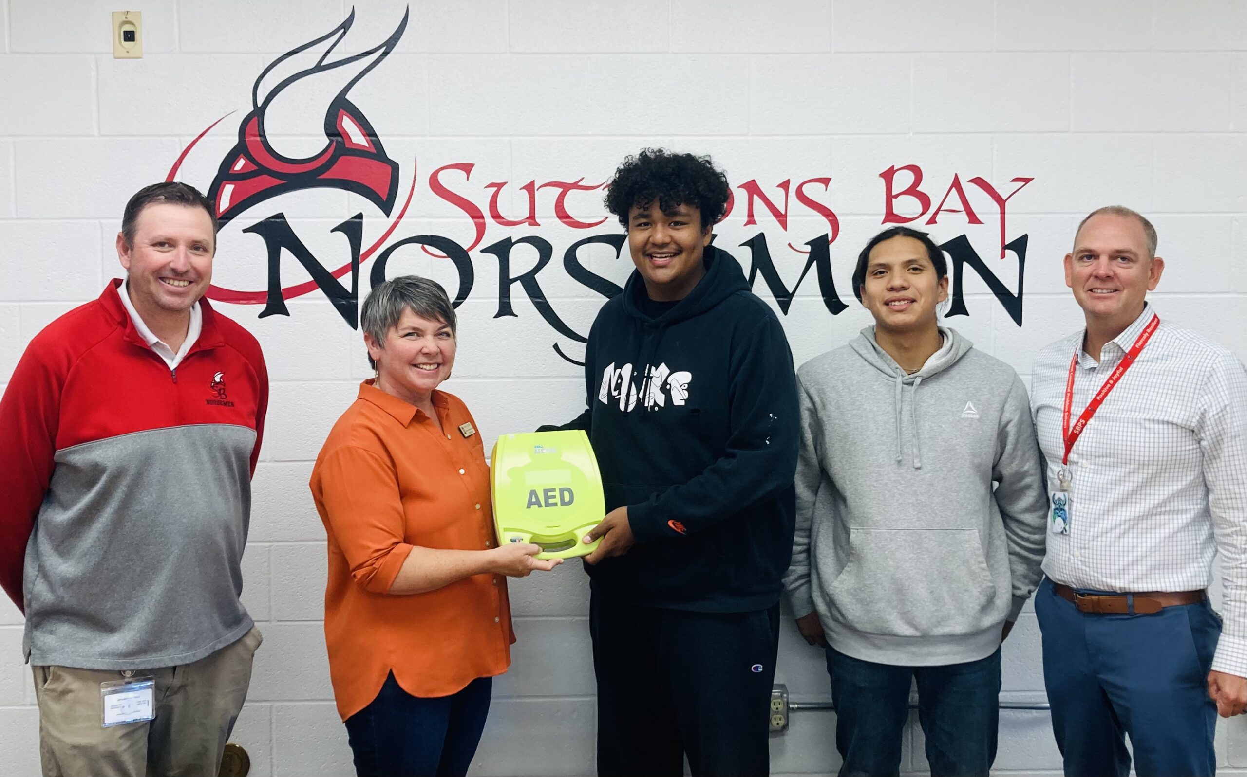 Michigan State Representative Betsy Coffia passing an automated external defibrillator to students Zachariah Raphael, Damien Chippewa, Superintendent Casey Petz and Chief Operations Officer Tim Smith in front of a Suttons Bay Norsemen mural at Suttons Bay High School.