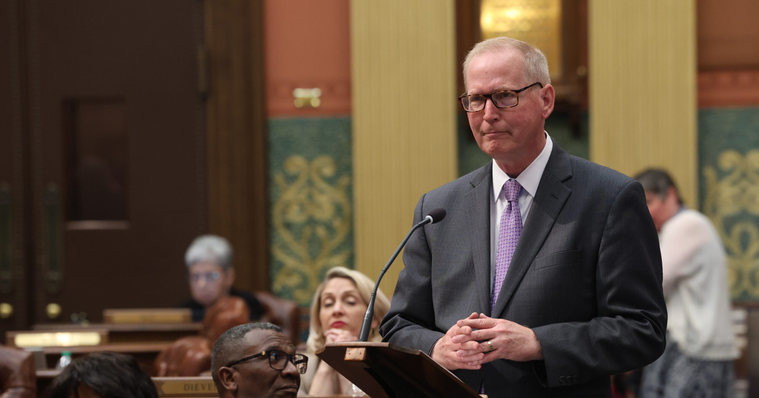 Michigan State Representative Jim Haadsma stands behind a podium on the House Floor to deliver a speech.