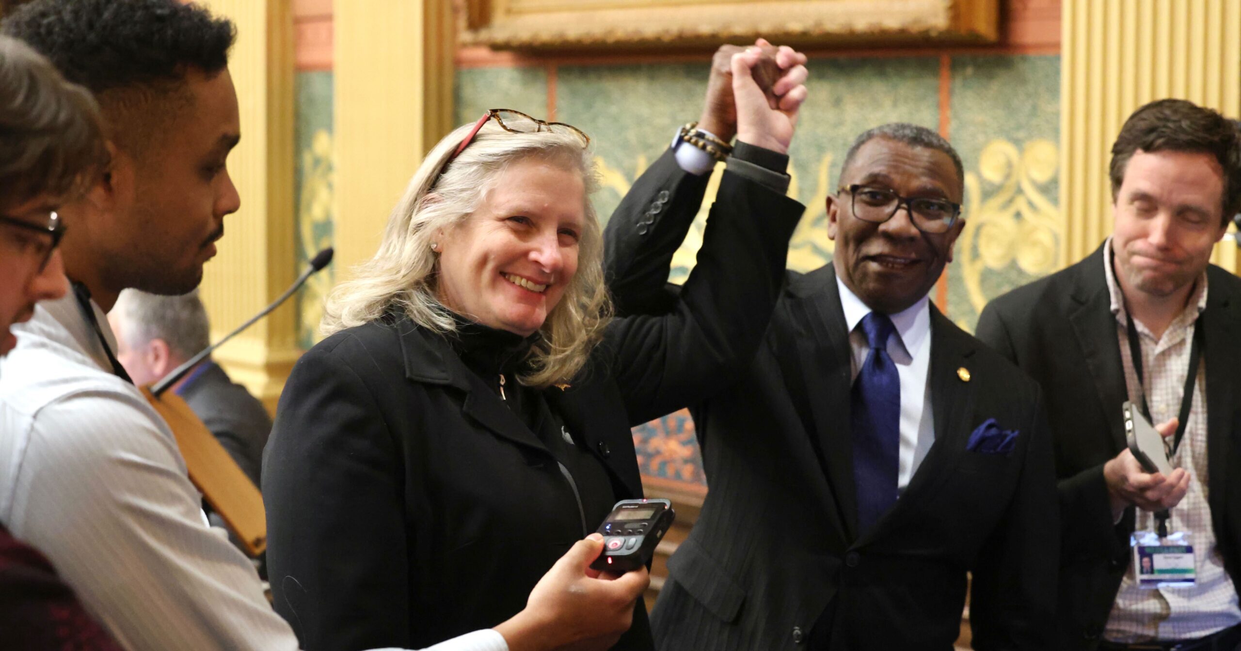 Michigan State Representatives Jenn Hill and Amos O'Neal smiling and holding hands above their heads while talking to reporters on the House floor.