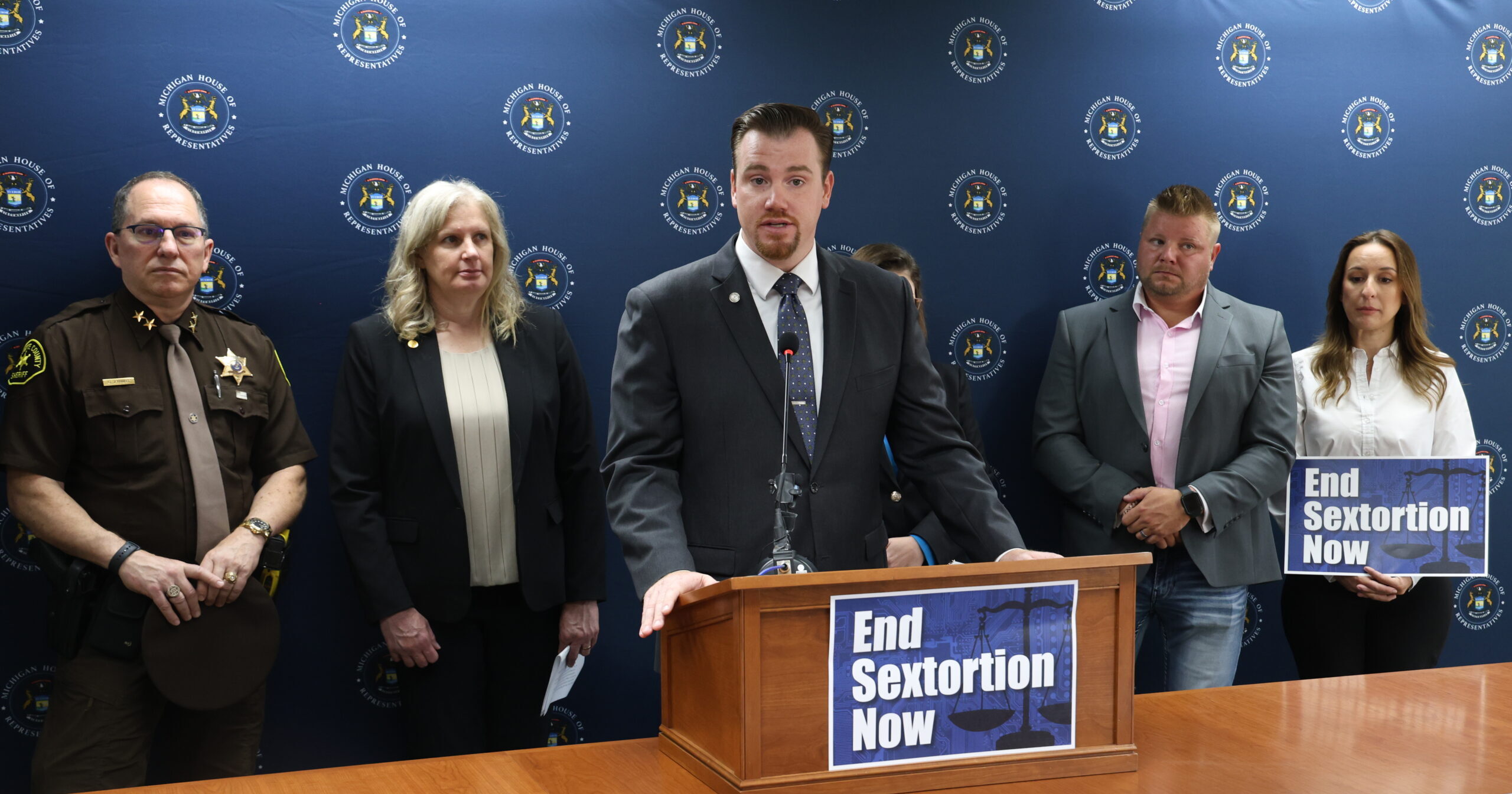 Democratic Floor Leader John Fitzgerald speaking behind a podium that reads "End Sextortion Now."