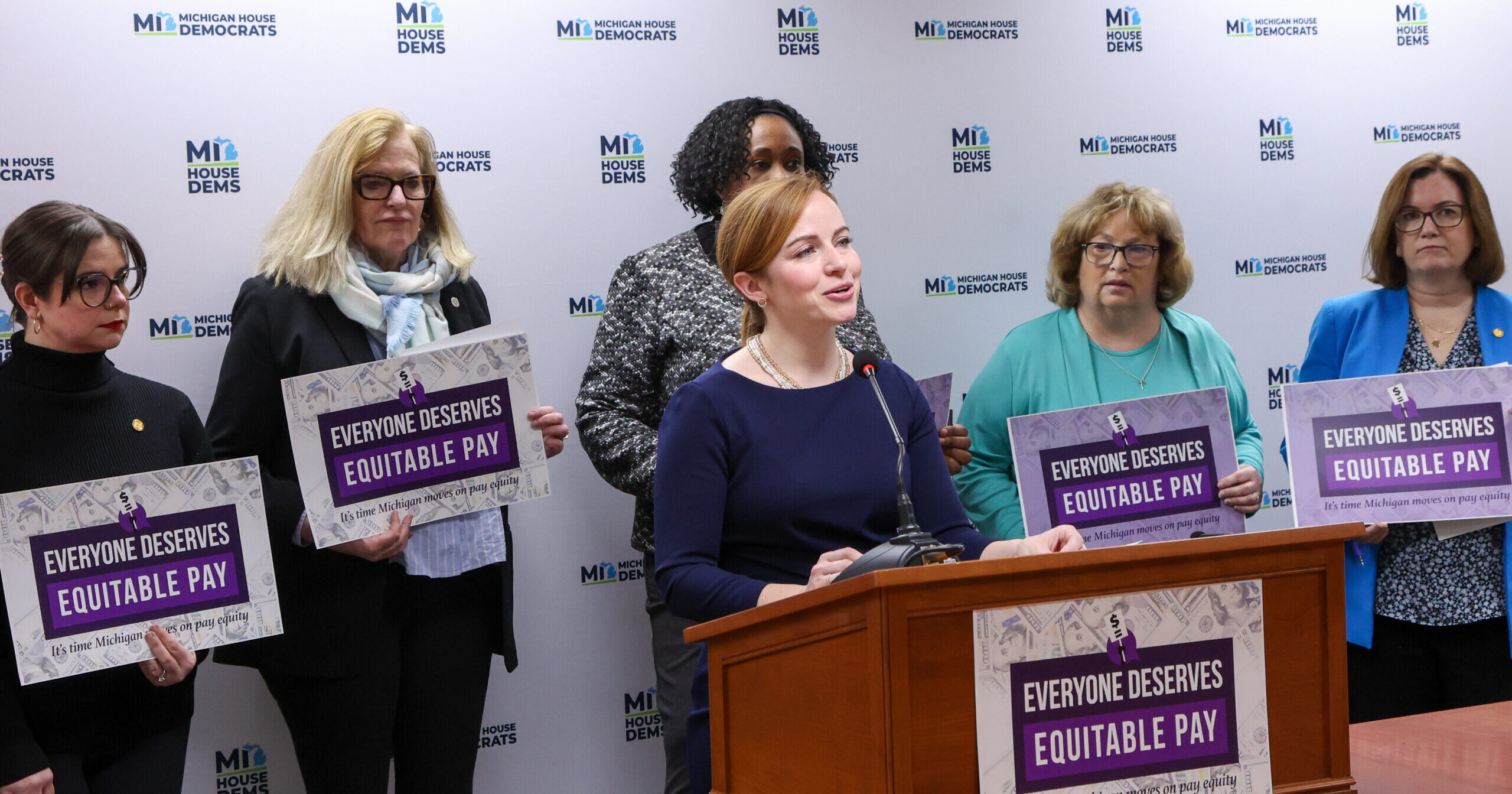 Michigan State Representative Erin Byrnes speaking behind a podium that reads "Everyone Deserves Equitable Pay." Members of the Progressive Women's Caucus stand behind her holding signs with the same message.