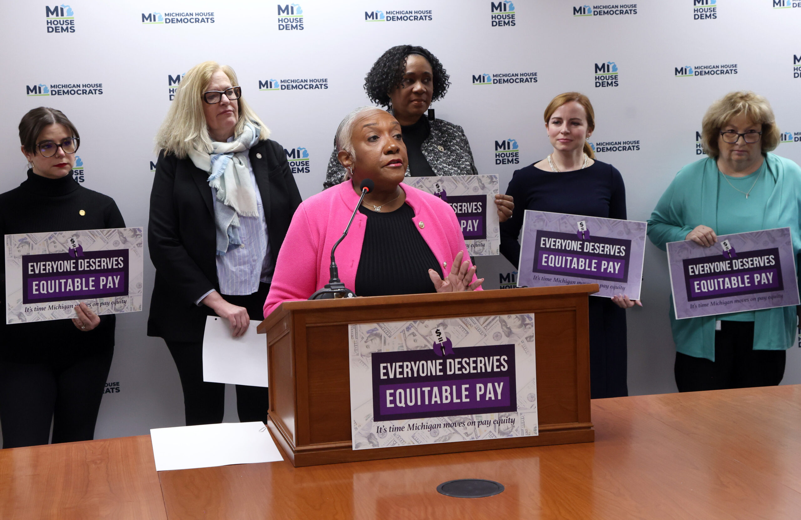 Michigan State Representative Helena Scott speaking behind a podium with a sign that reads "Everyone Deserves Equitable Pay." Members of the Progressive Women's Caucus stand behind her holding more signs.