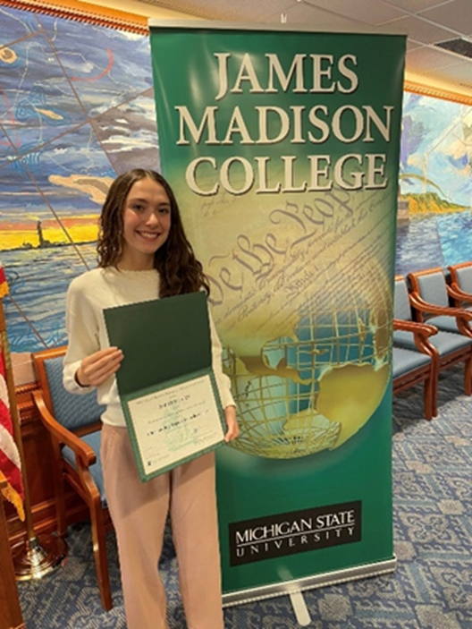 2025 Rosenthal Award winner Tabitha Garza posing with her award in front of a banner that reads "James Madison College, Michigan State University."