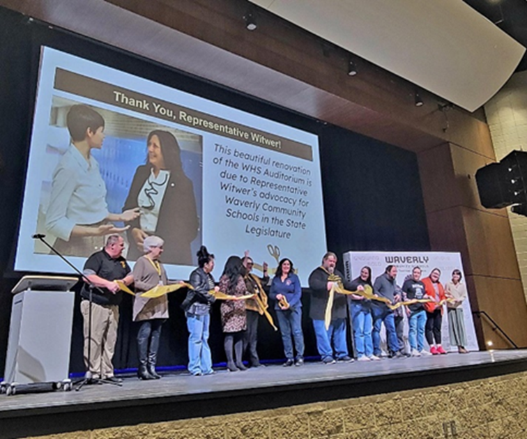 Michigan State Representative Angela Witwer and Waverly High School staff on stage cutting a ribbon in front of a presentation that reads "Thank you, Representative Witwer! This beautiful renovation of the WHS Auditorium is due to Representative Witwer's advocacy for Waverly Community Schools in the State Legislature."