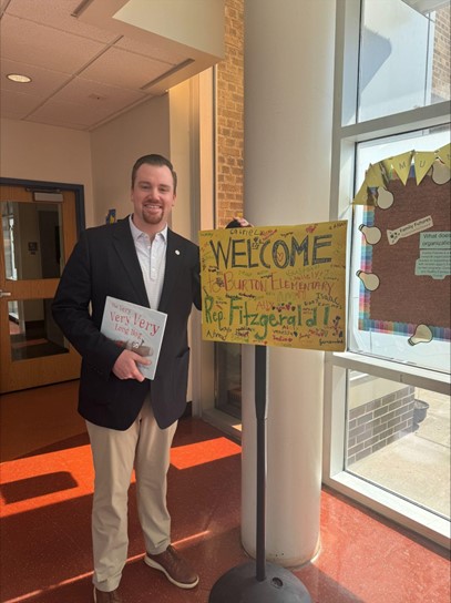A photo of Michigan state Representative John Fitzgerald holding a picture book and posing with a hand-made sign that reads "Welcome to Burton Elementary, Representative Fitzgerald!"