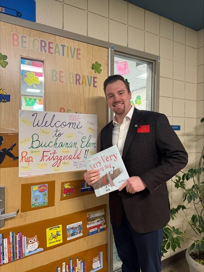 Michigan state Representative John Fitzgerald holding a picture book and posing for a photo with a hand-made sign that reads "Welcome to Buchanan Elementary, Representative Fitzgerald."