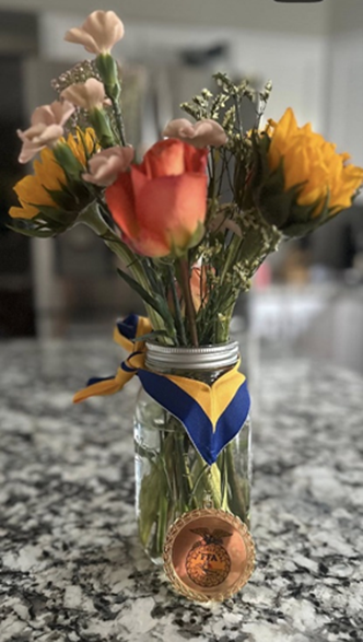 Red, yellow and pink cut flowers in a bouquet in a clear mason jar. Wrapped around the jar is a bronze medal from the Future Farmers of America.