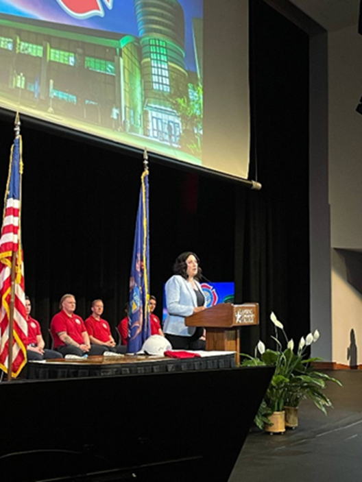 Michigan state Representative Angela Witwer speaking on stage in front of the graduating class of Lansing Community College's Fire Academy.
