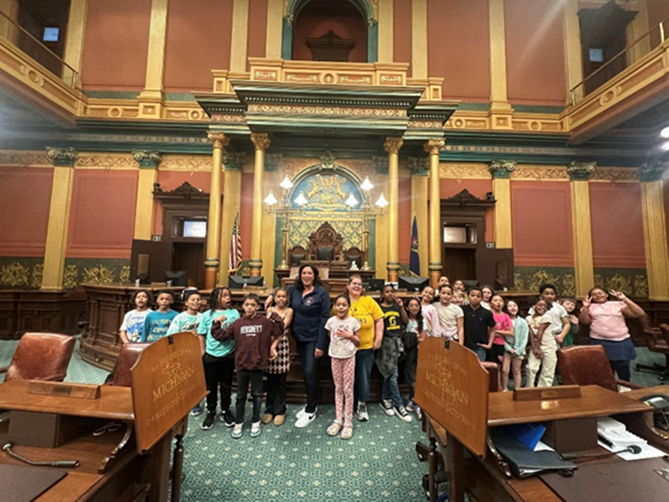 Michigan state Representative Angela Witwer posing with a class of elementary schoolers during a tour of the House floor in the Michigan Capitol Building.