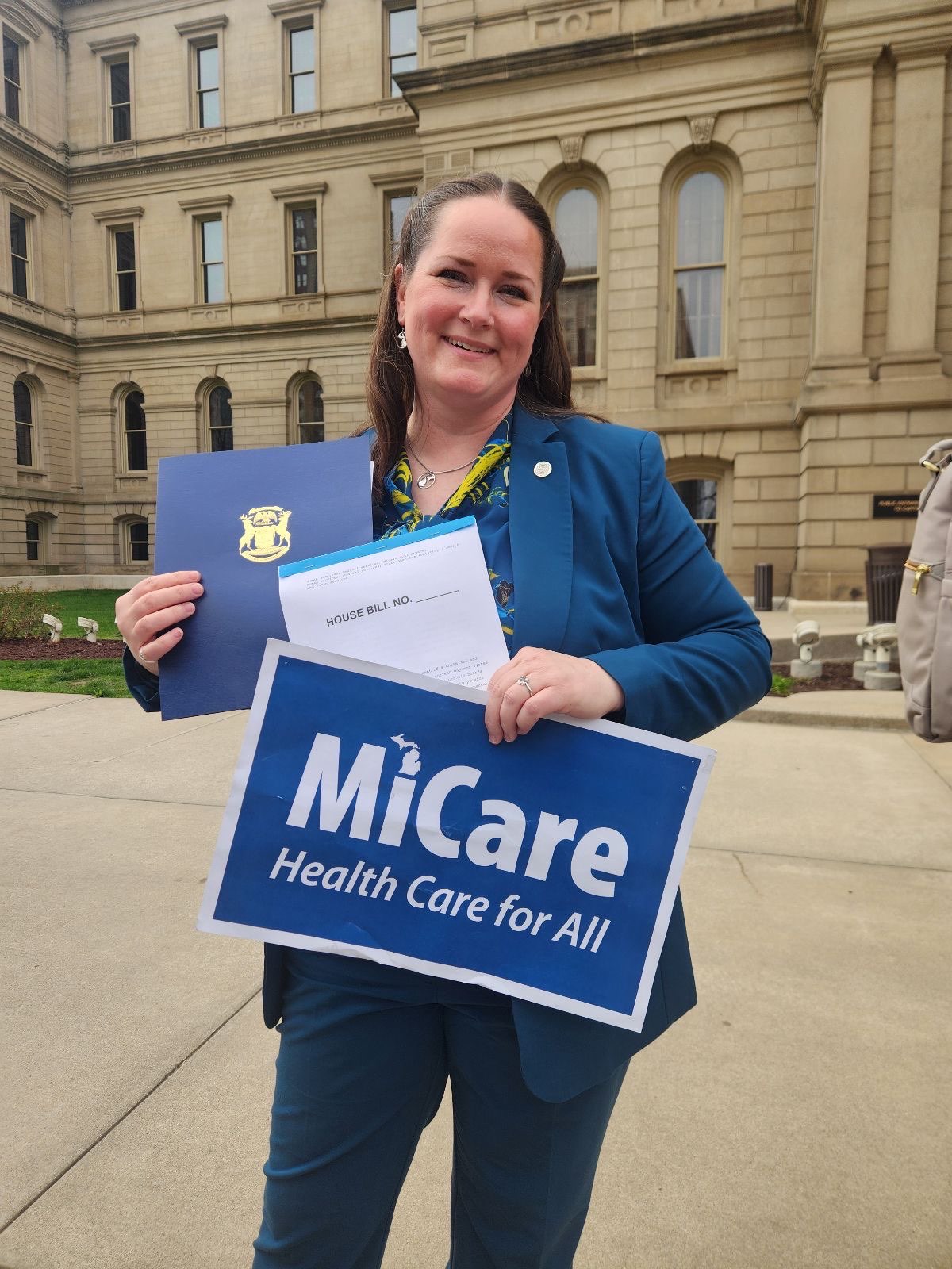 Michigan State Representative Carrie Rheingans stands in front of the Capitol with a sign that says "MiCare, Health Care for All."