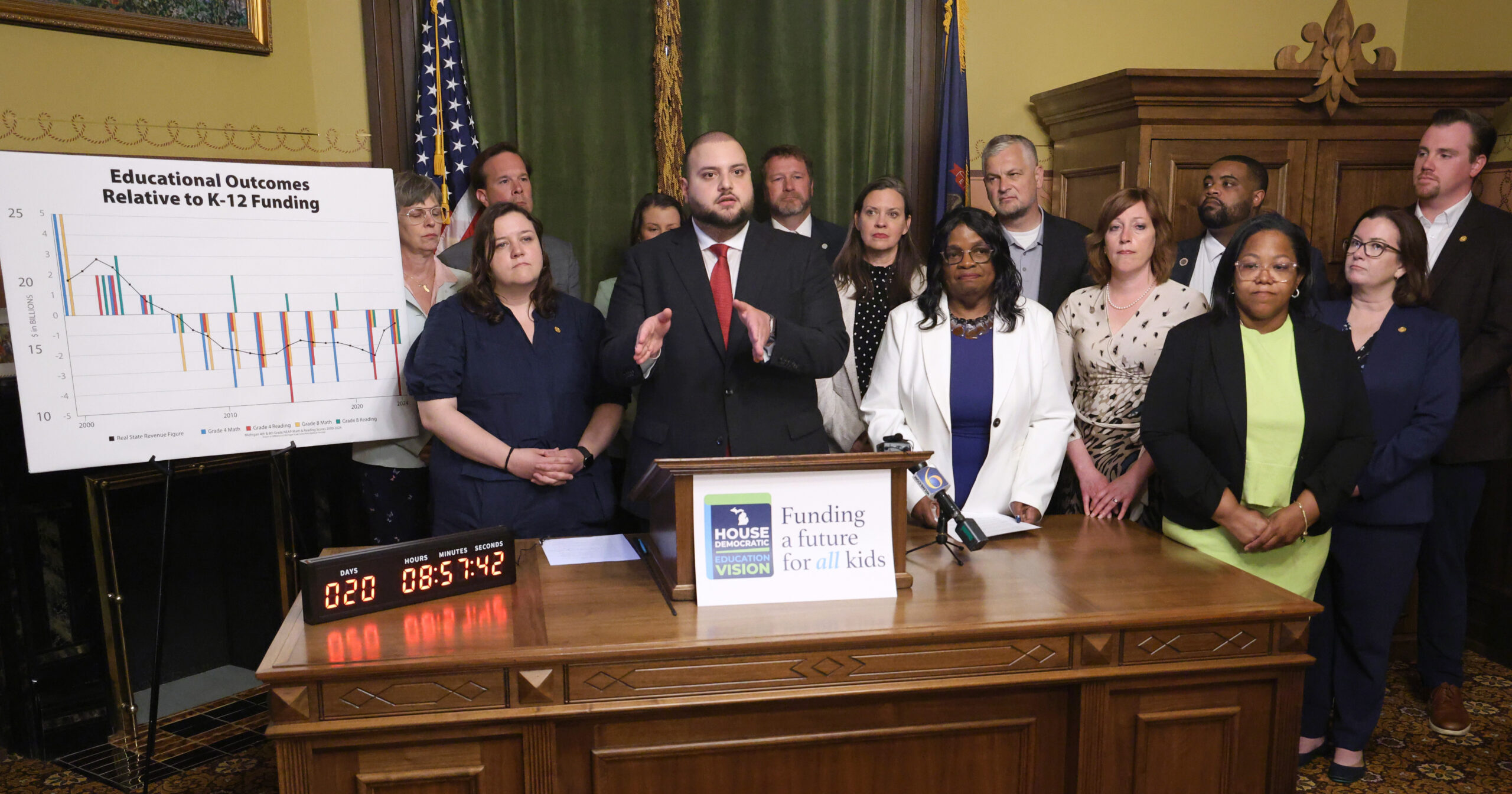 Rep. Alabas Farhat speaks at a podium in front of many House Democrats members.