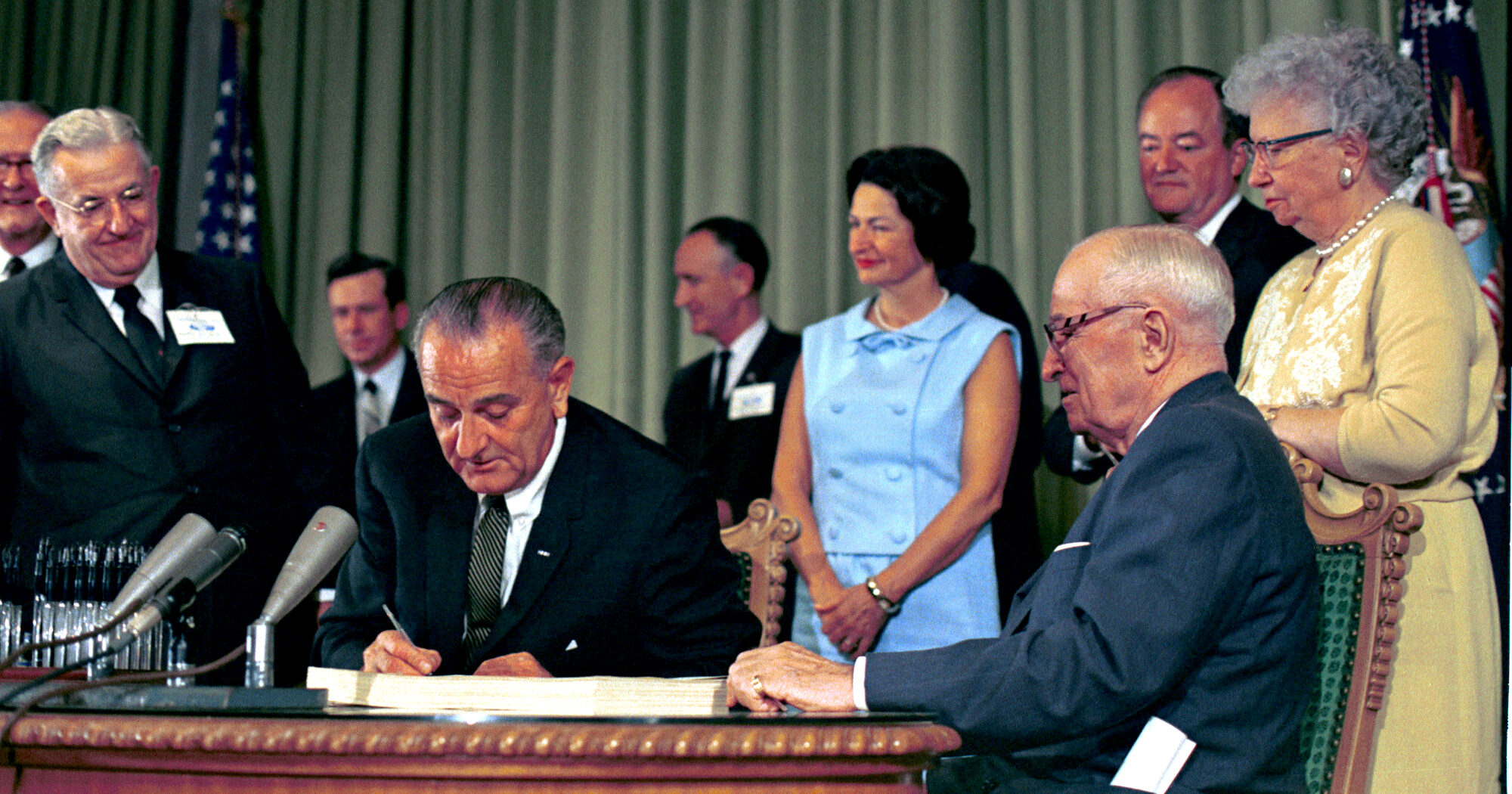 Historical photograph of President Lyndon B. Johnson signing the landmark Medicare and Medicaid Act into law on July 30, 1965, in Independence, Missouri, with former President Harry S. Truman at his side.