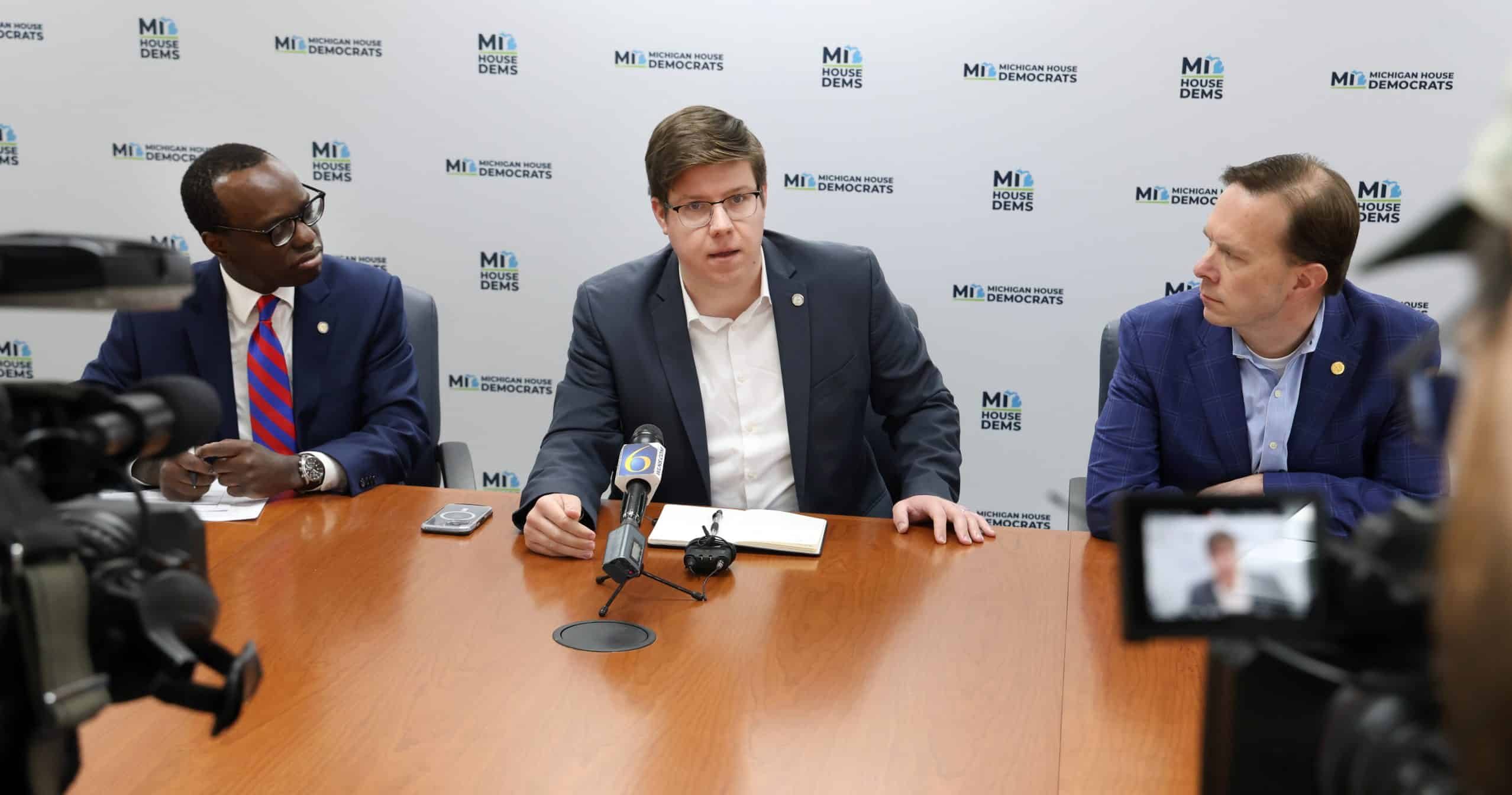 State Representatives Stephen Wooden and Matt Koleszar speaking at a press conference in front of a backdrop with the Michigan House Democrats logo.