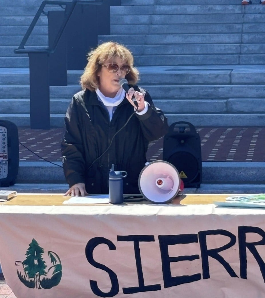 Michigan State Representative Reggie Miller speaking into a microphone while standing behind a table with a banner that reads "Sierra Club."