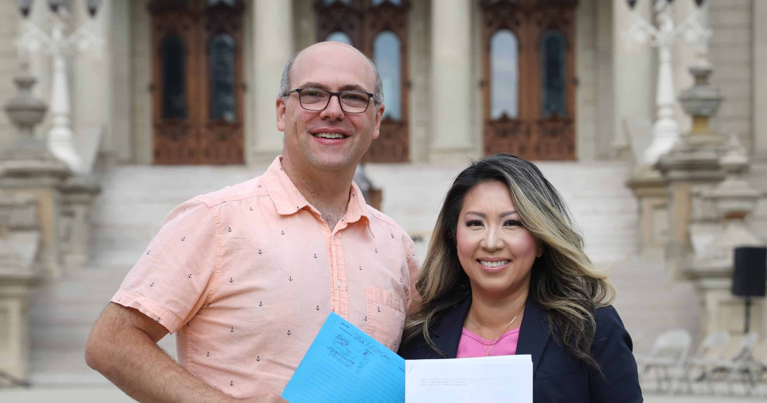 State Senator John Cherry and state Representative Mai Xiong holding paper copies of their legislation for a photo on the steps of the Michigan Capitol Building.
