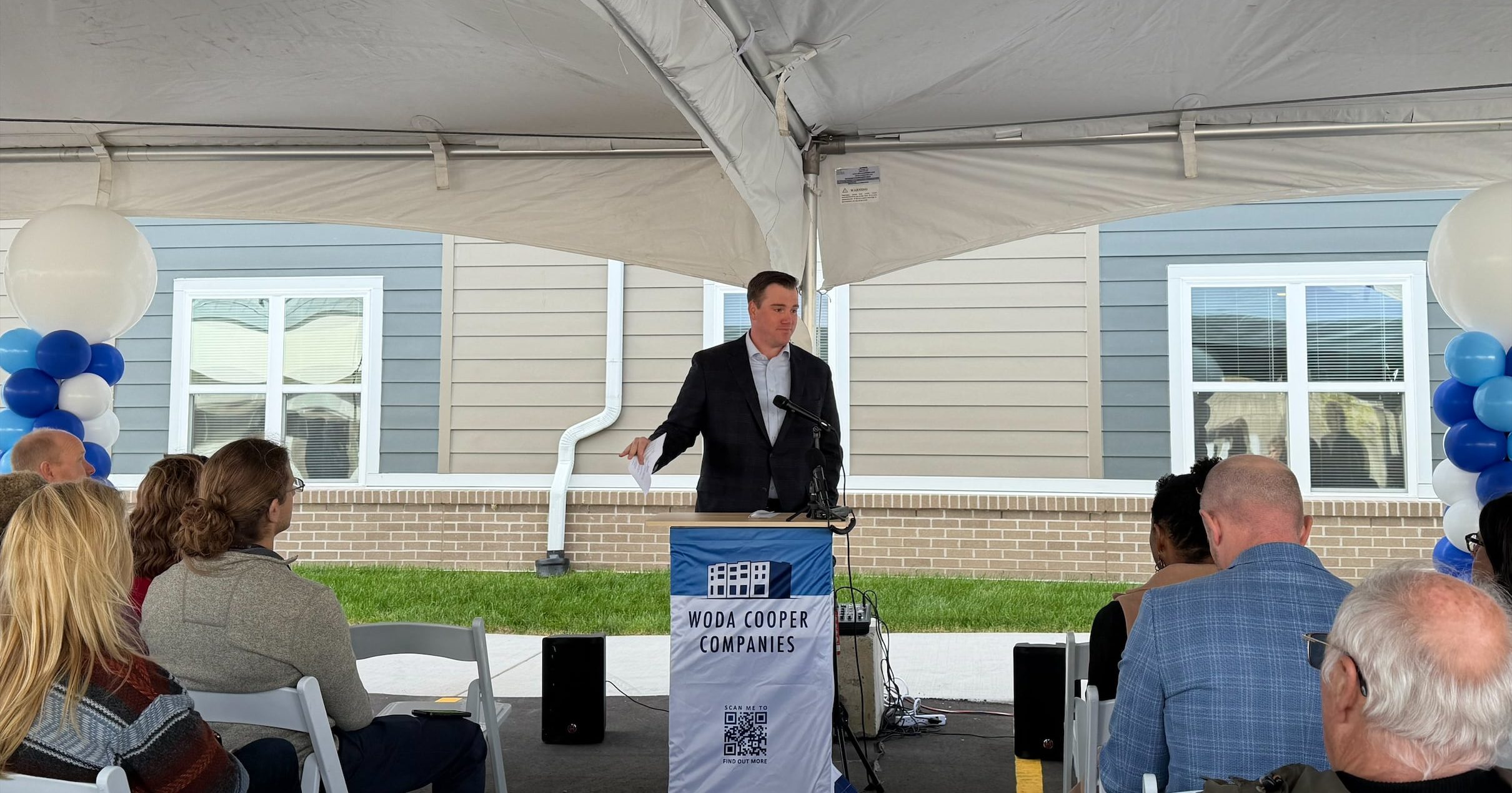 State Representative John Fitzgerald speaking to a crowd of community members in front of the Shea Ravines affordable housing development. Standing behind a podium and between blue and white balloons, Representative Fitzgerald speaks to the grand opening of Shea Ravines.